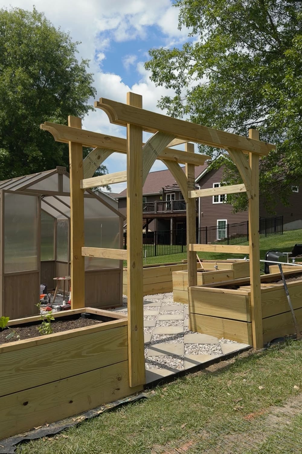Wooden DIY garden trellis arbor entrance with decorative curved corbels, framing a checkered paver and gravel walkway between raised garden beds with a greenhouse in the background