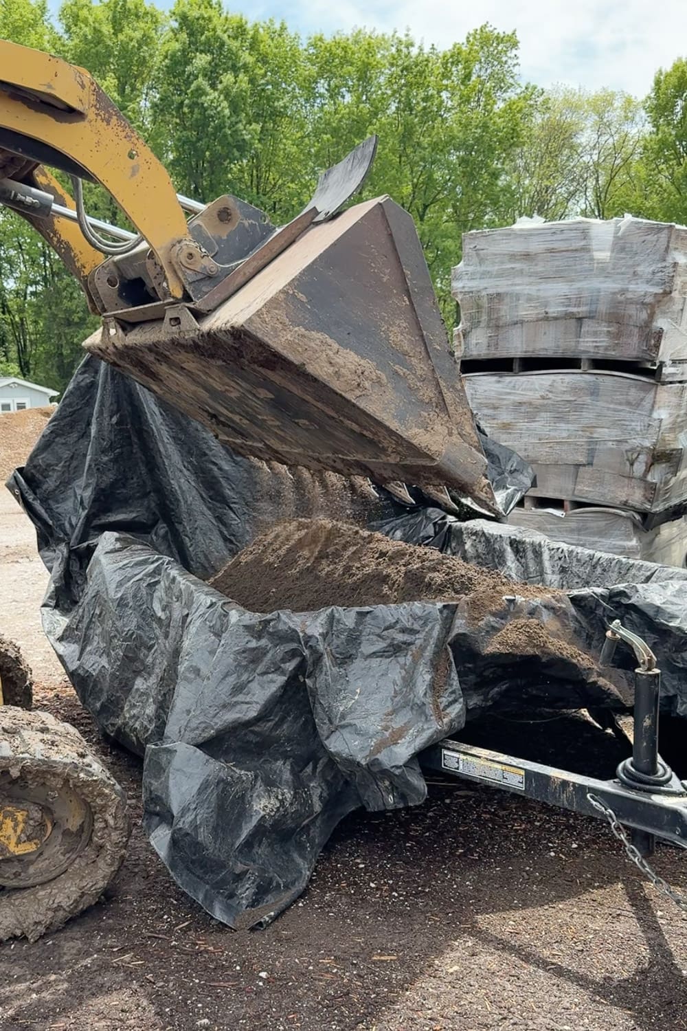 Skid steer loader scooping bulk soil into a tarp-lined utility trailer at a landscape supply yard, with pallets of mulch bags stacked nearby.