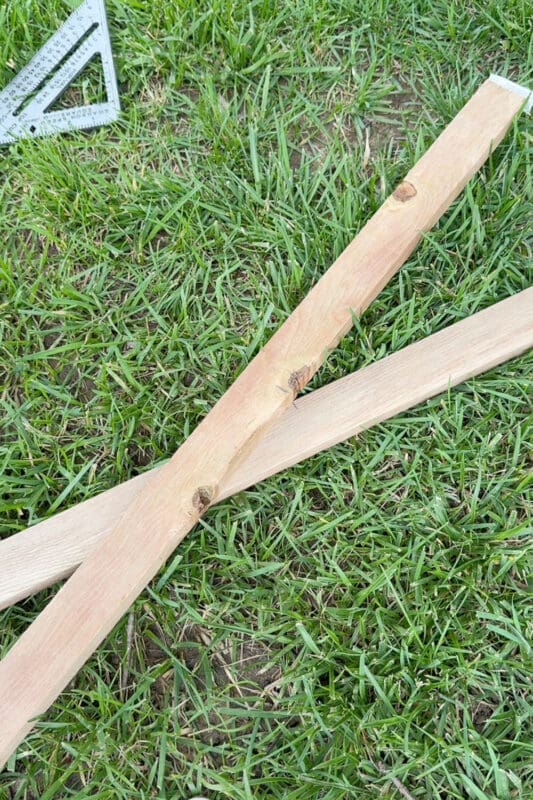 Two wood stakes crossed to form an X for a budget friendly, wood climbing support, with a speed square laid on the grass for marking the angle