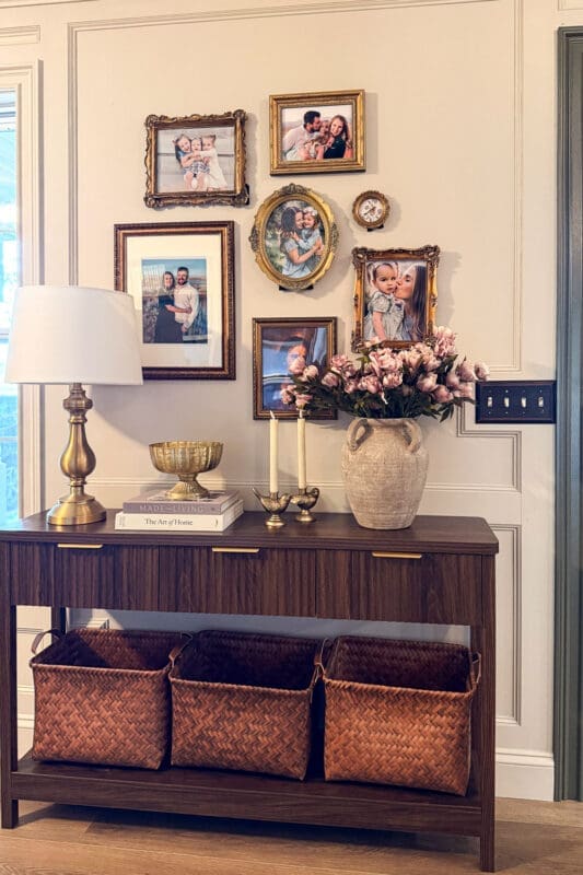 Entryway decor with a dark wood console table, brass lamp, stone vase of pink peonies, gold gallery wall, and woven storage baskets