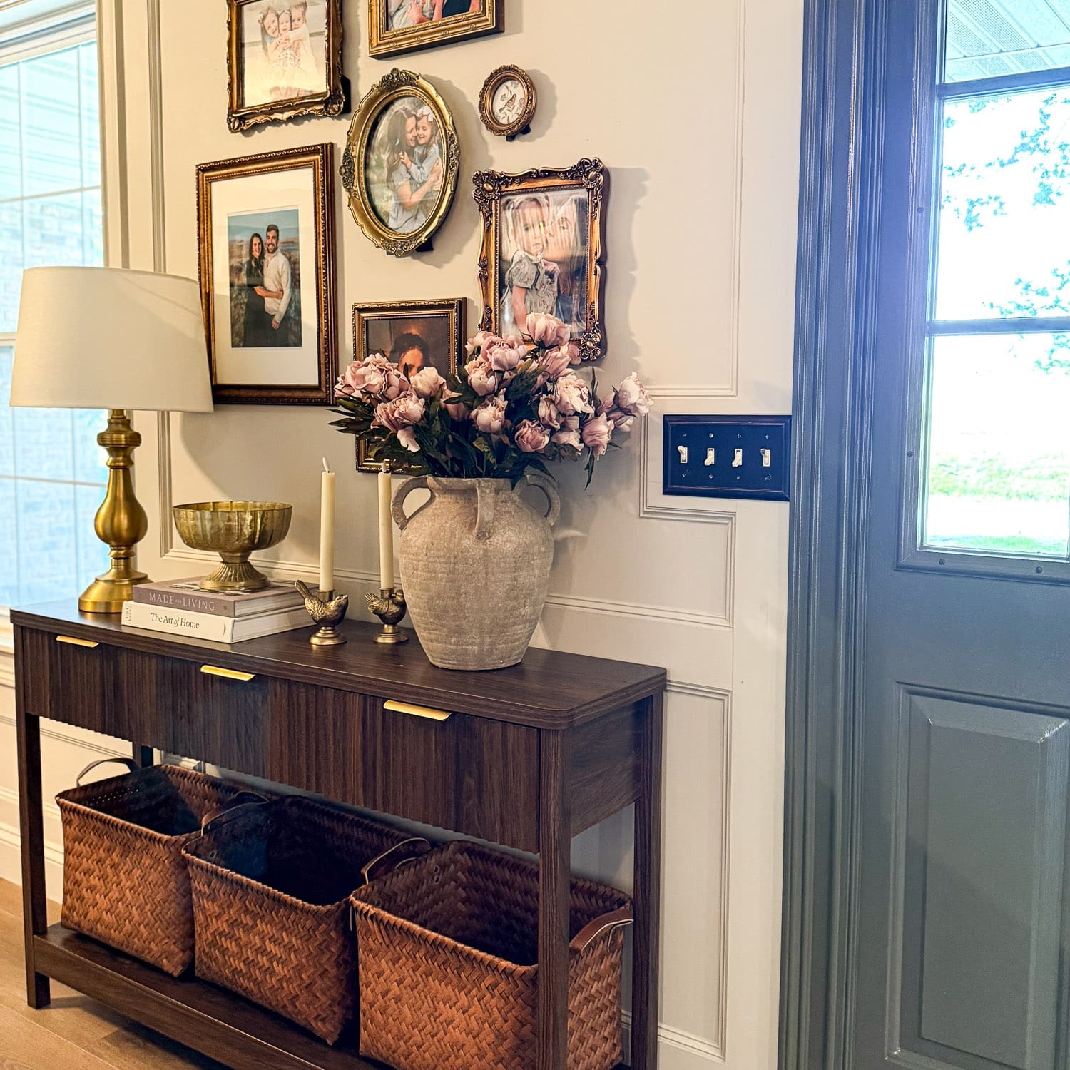 Entryway decor featuring a dark wood entry table styled with brass lamp, vintage stone vase with pink peonies, and a gold-framed family photo gallery wall