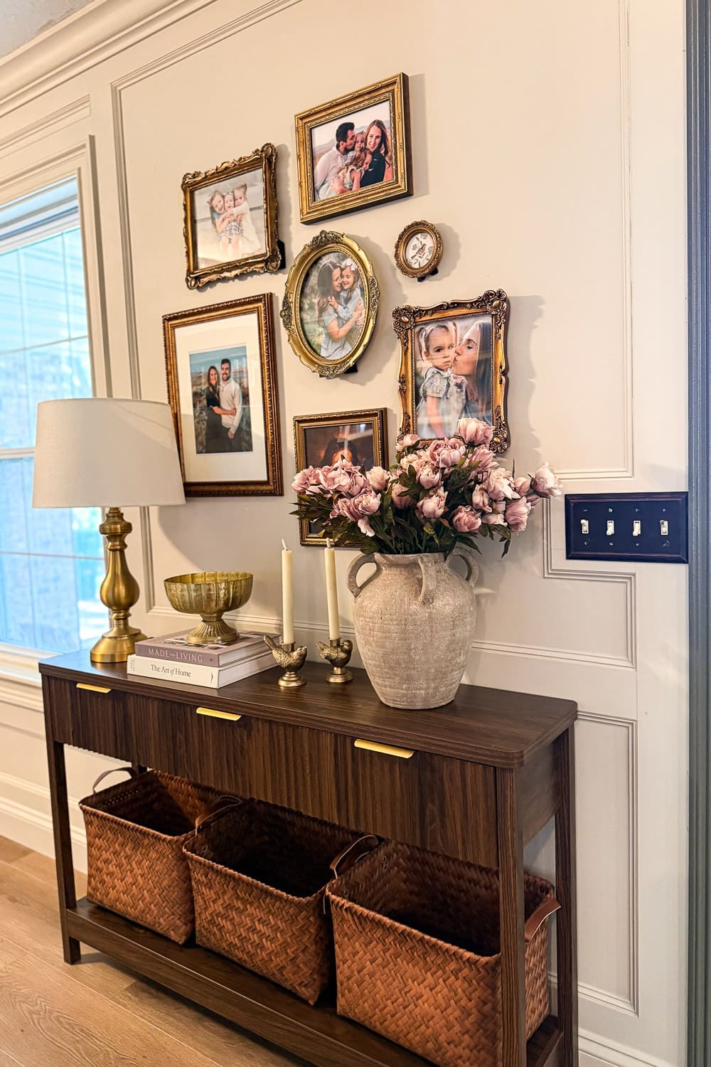 Entryway decor with a gold-framed family gallery wall, brass lamp, stone vase of pink peonies, and woven baskets on a dark wood console