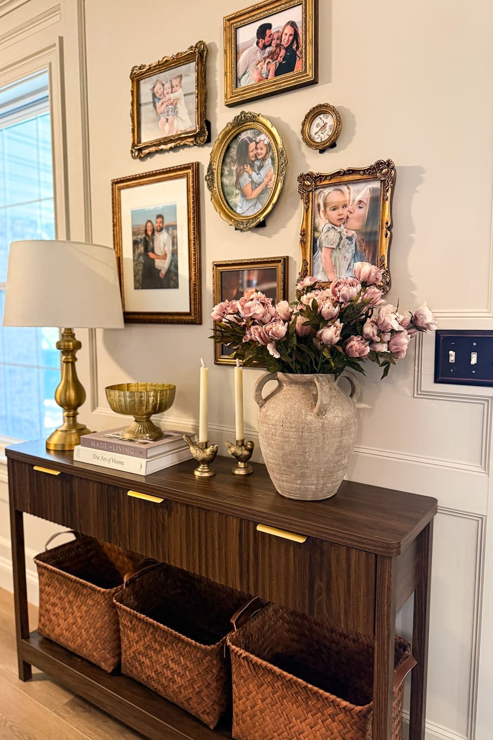 Entryway table decor with brass lamp, candlestick birds, stone vase of pink peonies, and woven baskets below dark wood console table with gold gallery wall
