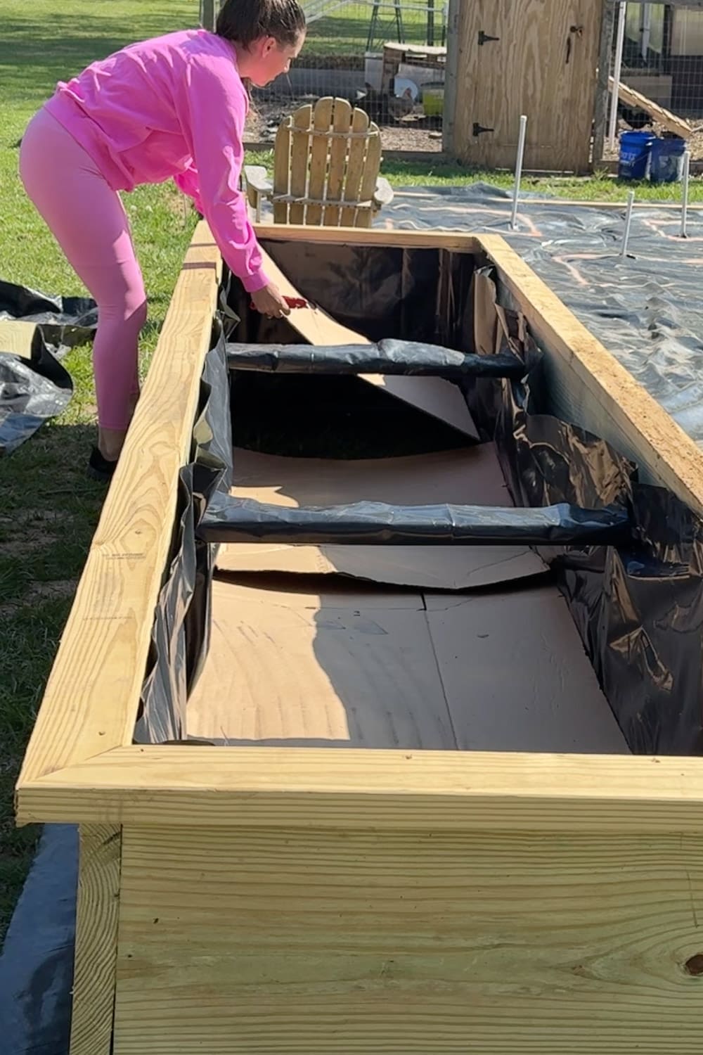 Woman in a pink sweatsuit lining a large wooden raised garden bed with black plastic sheeting and cardboard along the bottom and sides, the first step in how to fill a raised garden bed using the layering method.