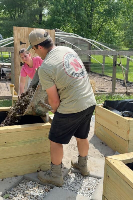 Husband in a Nebraska Huskers t-shirt dumping a wheelbarrow full of soil and organic matter into a wooden raised garden bed while his wife steadies the load — a teamwork moment showing how to fill a raised garden bed efficiently with bulk material.