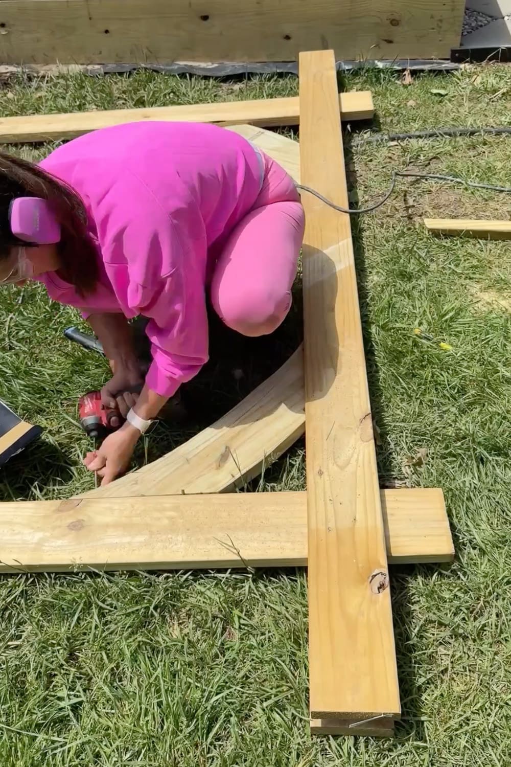 Woman in pink kneeling on the grass using a cordless drill to attach a curved corbel brace to the upright post of a DIY garden trellis arbor frame