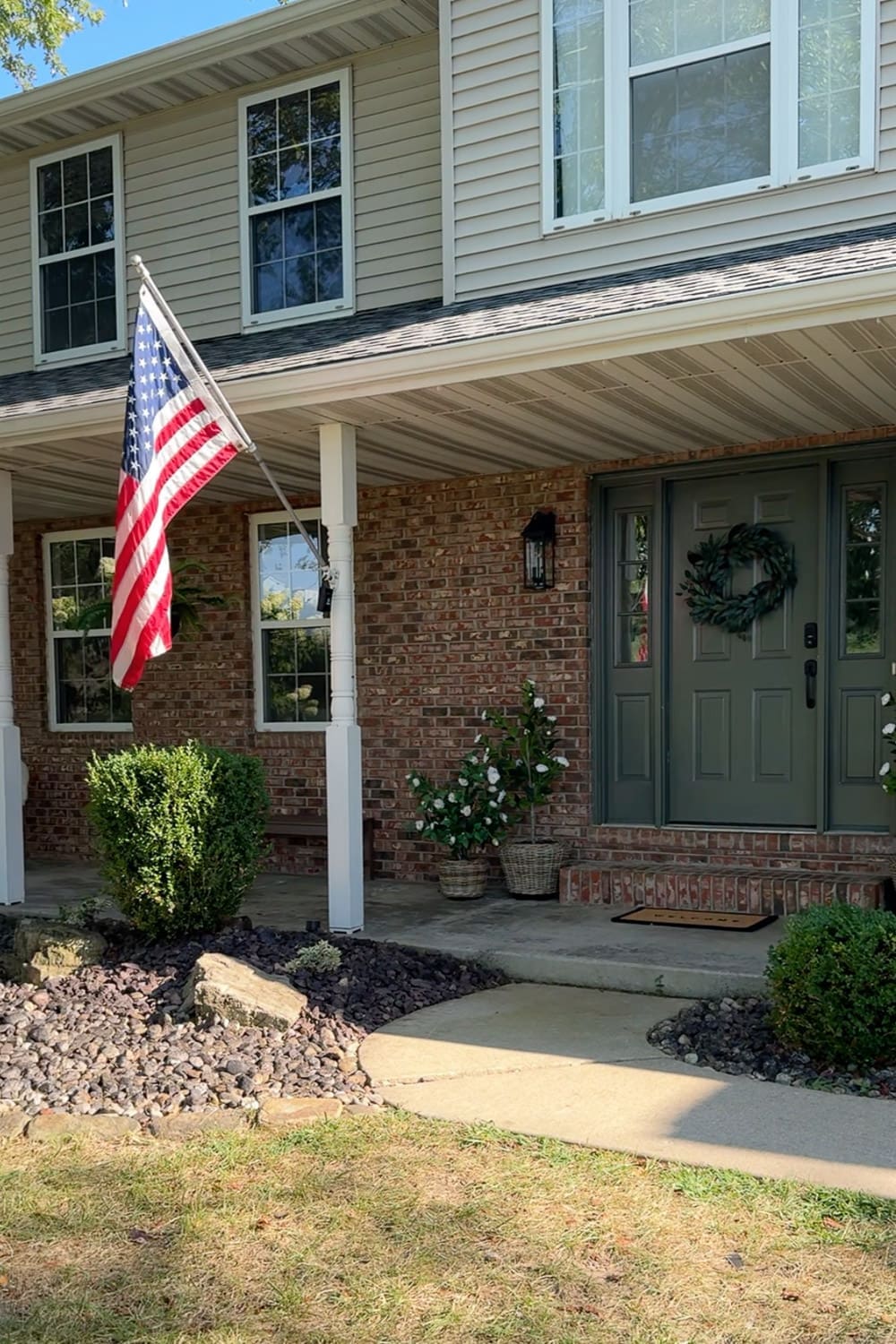American flag hanging from a brick front porch with a sage green front door, magnolia wreath, and white potted flowers