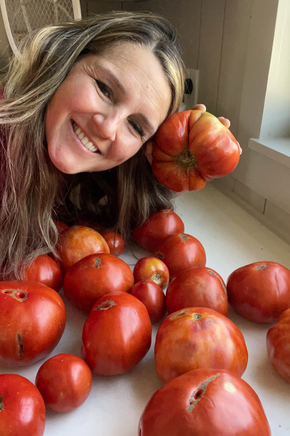 Smiling woman holding a homegrown heirloom tomato next to a fresh harvest from her summer tomato plants