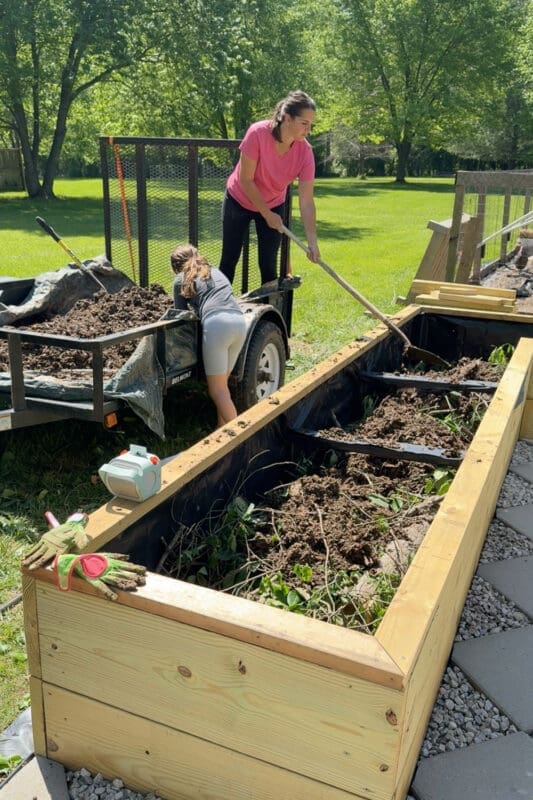 Mom in a pink shirt shoveling aged compost into a long wooden raised garden bed already layered with sticks, leafy greens, and yard waste, while her daughter helps unload more material from a utility trailer — showing the layering method for filling a raised garden bed on a budget.