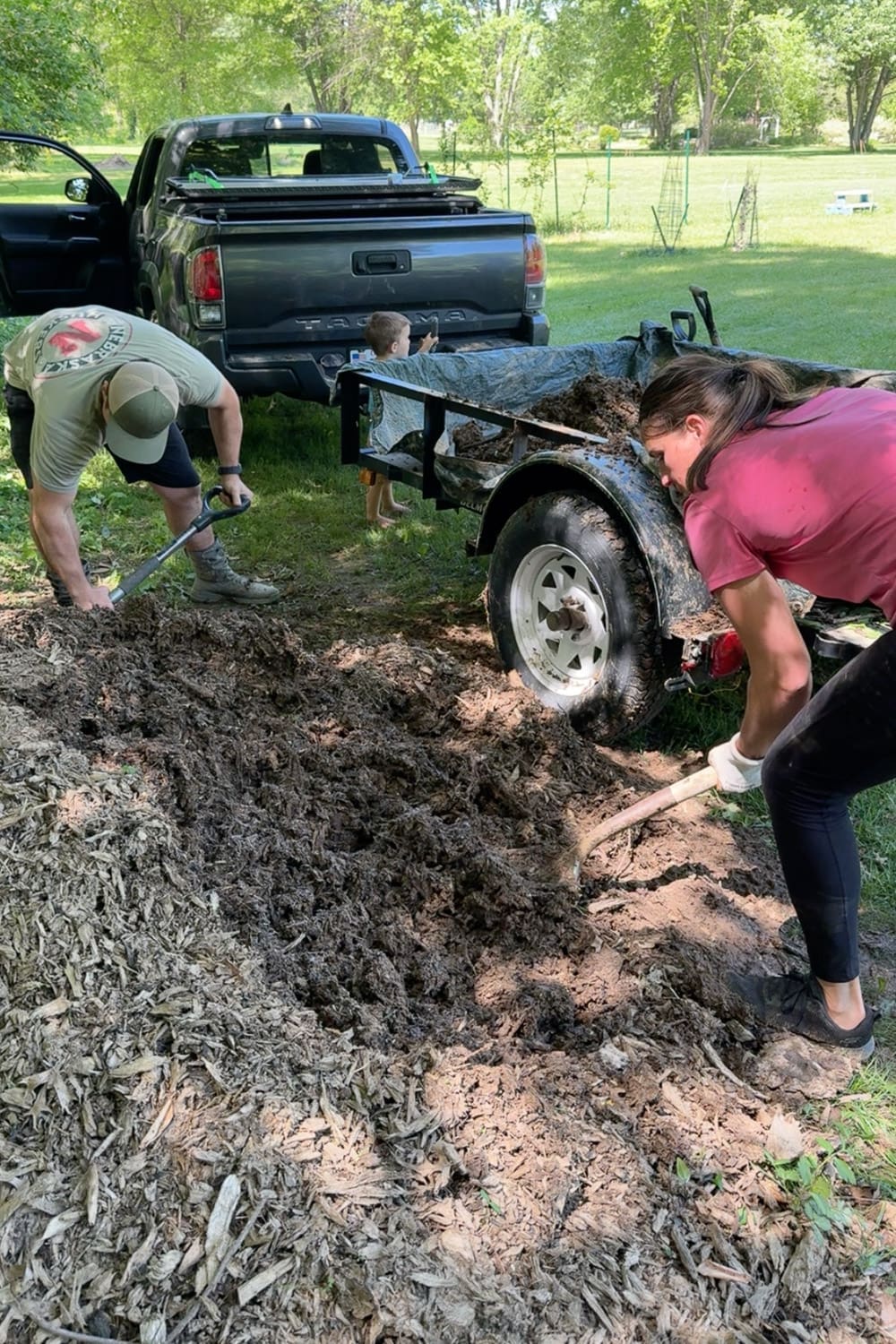 Husband and wife shoveling a large pile of aged wood chips and partially composted mulch into a utility trailer behind a Toyota Tacoma, gathering organic material for the soil layers in a raised garden bed, with a young child helping in the background.