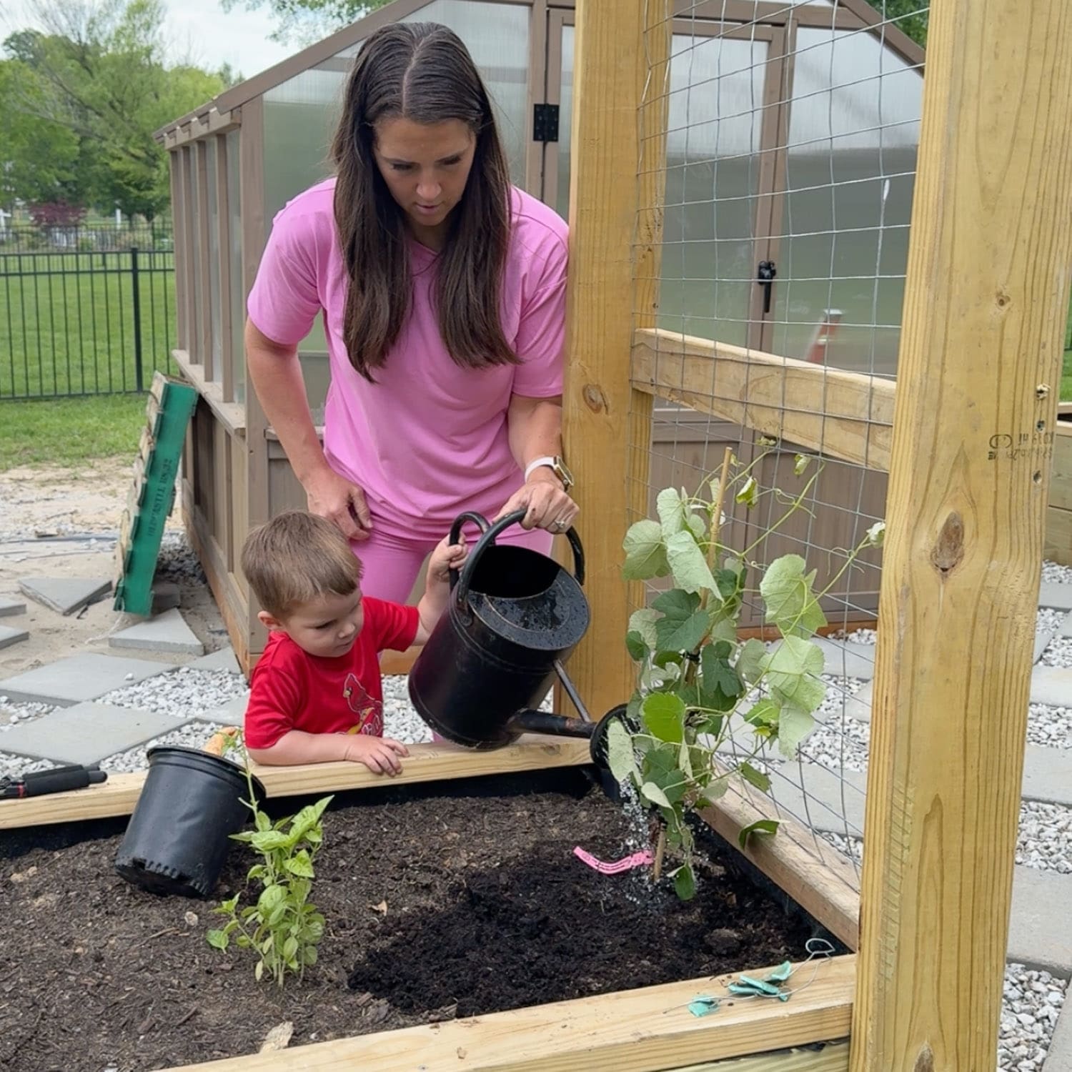 Mom and young son watering newly planted vegetables in a raised bed vegetable garden with a trellis and greenhouse in the background