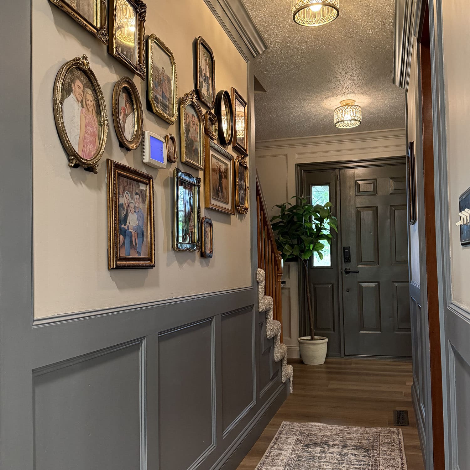 Narrow hallway gallery wall with mismatched gold frames above gray picture frame moulding, leading to a dark moody front door