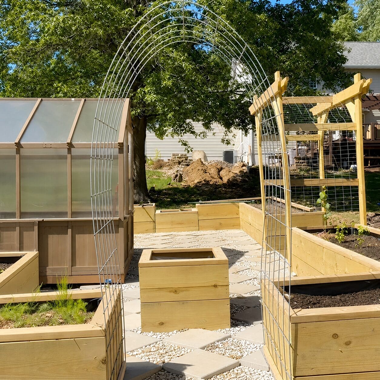 Arched DIY garden trellis made from wire panels over a gravel path between wooden raised garden beds, with a greenhouse and pergola in the background