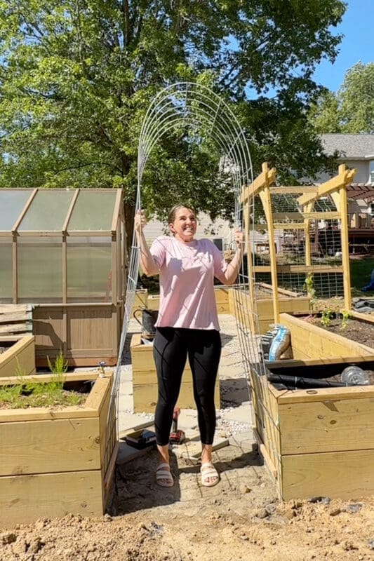 Woman standing under a finished arched garden trellis built over the path between two raised garden beds in a backyard vegetable garden