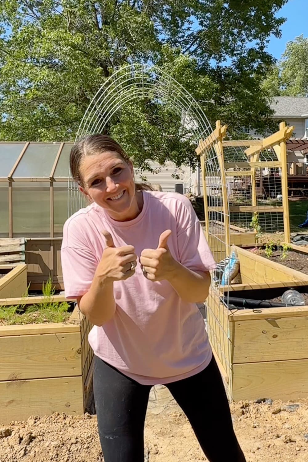 Woman giving two thumbs up in front of a completed arched garden trellis between raised garden beds in a backyard