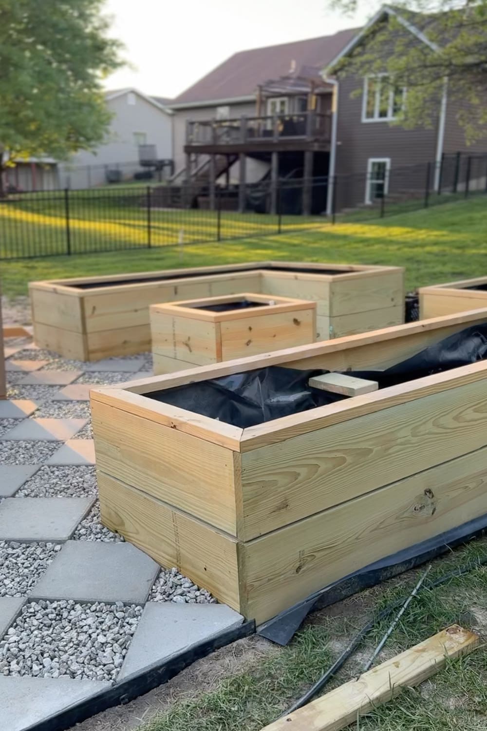 Freshly built wooden raised garden beds lined with landscape fabric, arranged around a checkerboard gravel and paver pathway in a backyard