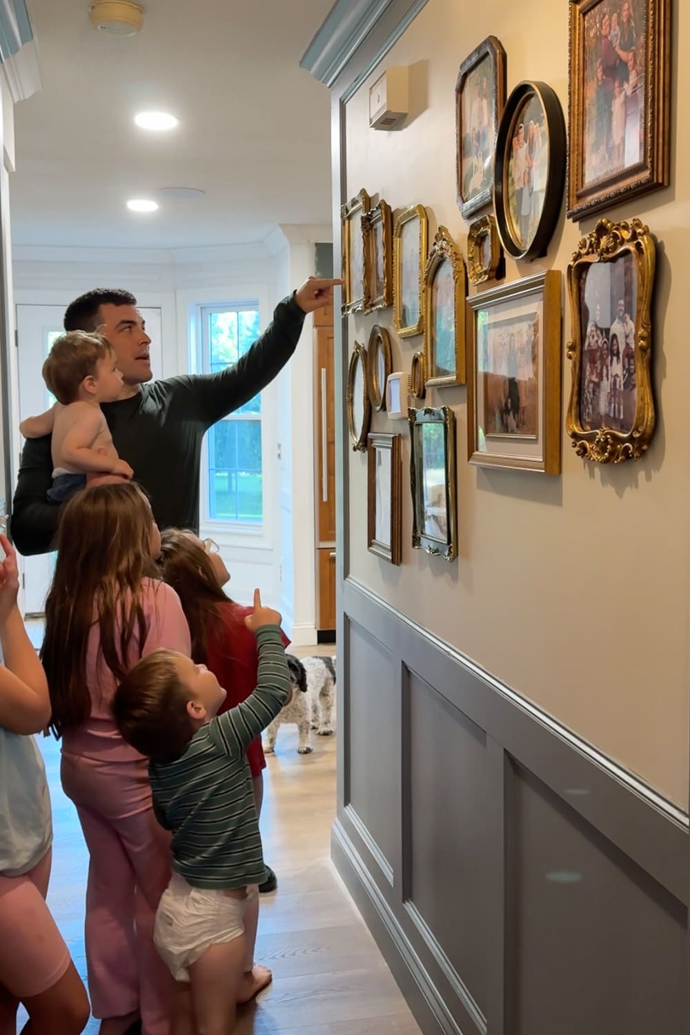 A dad holding a baby and three young children pointing up at the finished hallway gallery wall of mismatched gold frames