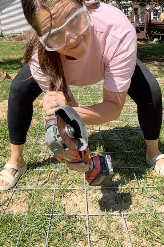 Woman wearing safety glasses using a cordless angle grinder to cut a metal cattle panel to size for a DIY garden trellis