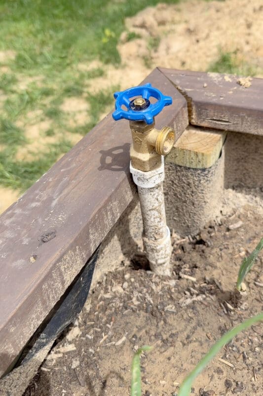 Brass spigot with a blue valve handle mounted at the corner of a wooden raised garden bed for DIY irrigation watering system