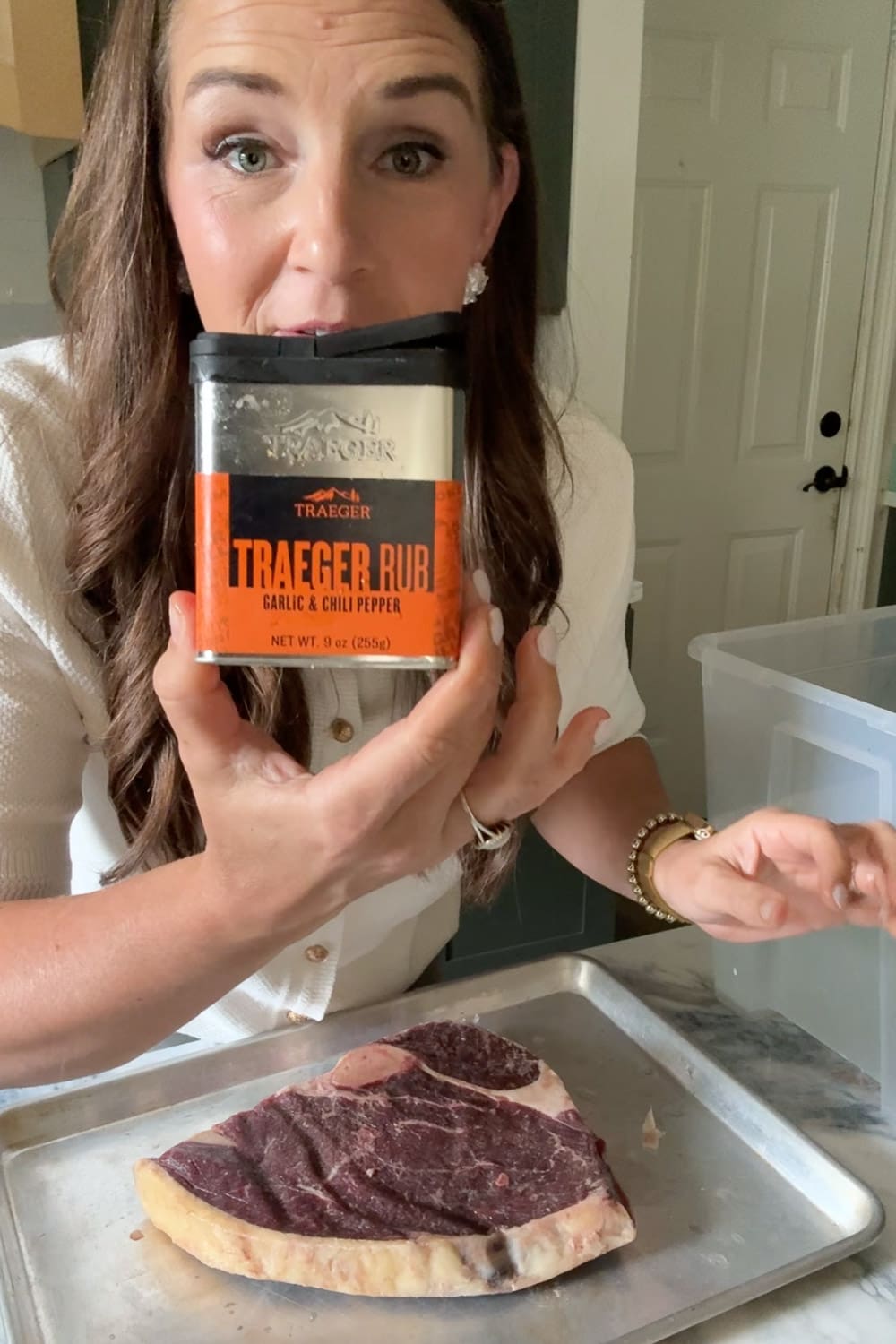 Holding a jar of Traeger garlic and chili pepper rub above a raw bone-in steak before sous vide cooking