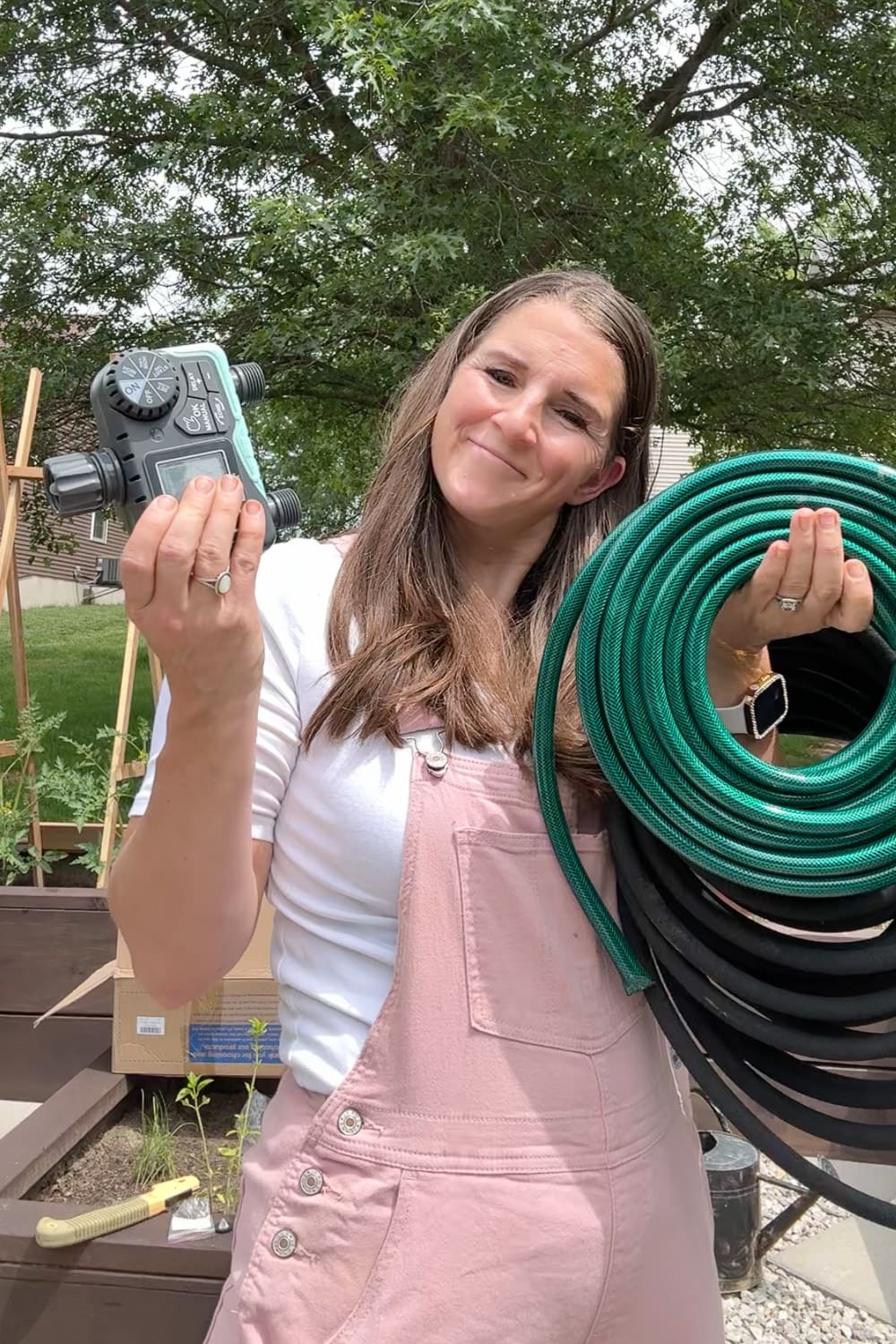 Crystel holding a digital water timer and coiled green and black soaker hoses, the supplies needed to set up automatic watering for raised garden beds