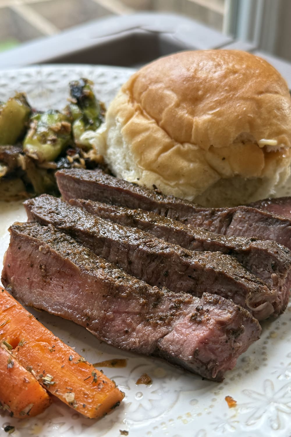 Sliced sous vide steak on a white plate with a dinner roll, roasted carrots, and brussels sprouts