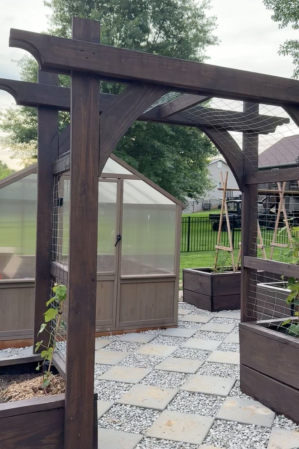 Finished dark stained raised garden beds and pergola archway framing a greenhouse and paver path