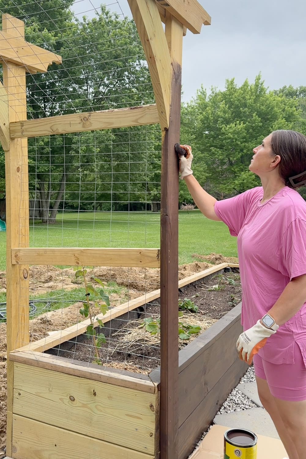 Woman hand-staining a pergola post above a raised garden bed with a sponge