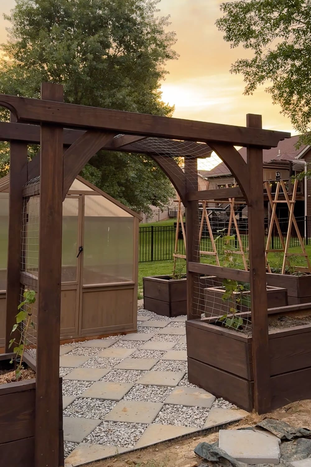 Dark stained raised garden beds and pergola archway at sunset with greenhouse and paver path