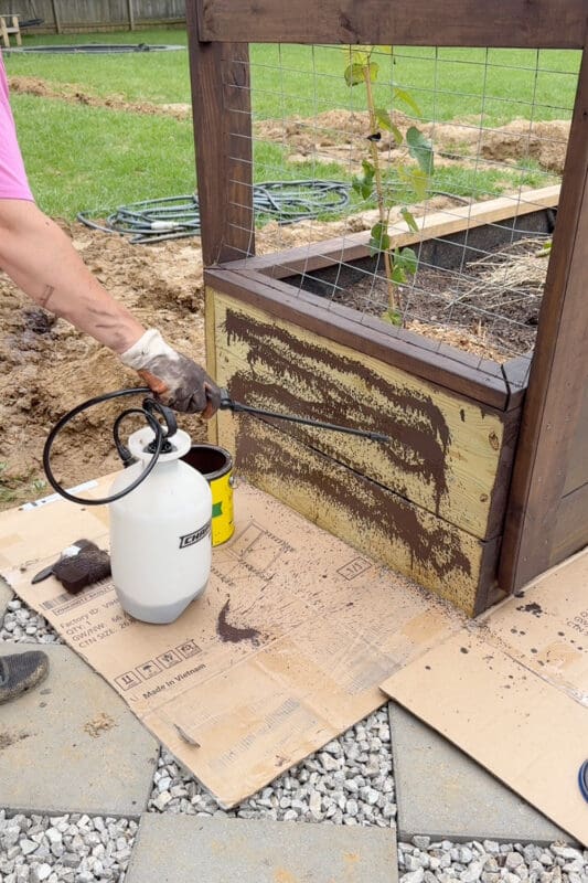 Using a pump sprayer to apply stain to a raised garden bed over a cardboard drop cloth