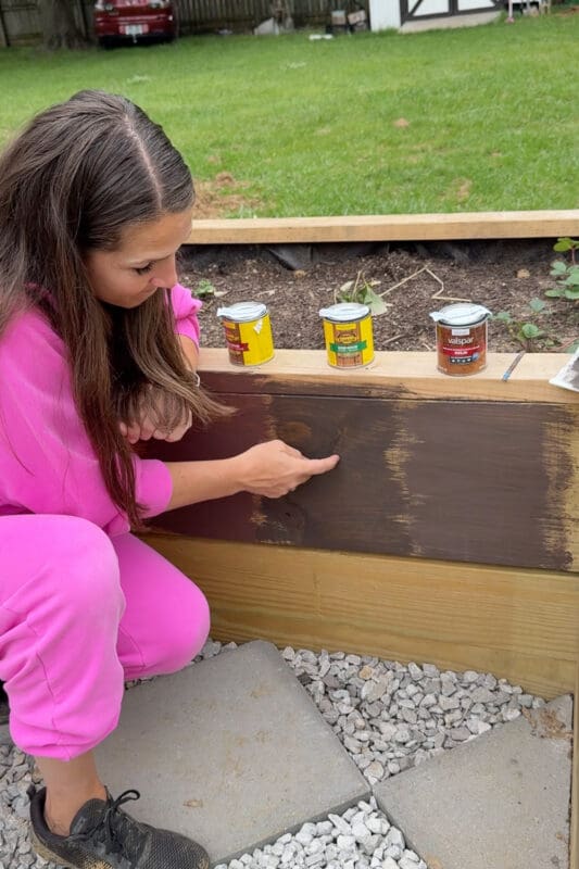 Woman comparing stain color swatches on a pressure treated raised garden bed before staining