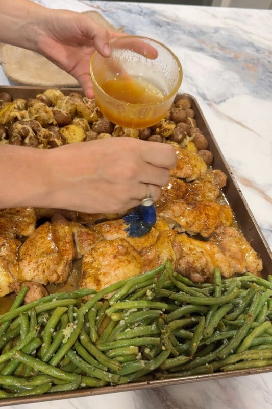 Crystel brushing honey glaze over roasted chicken thighs on the sheet pan, with parmesan potatoes and garlicky green beans alongside.