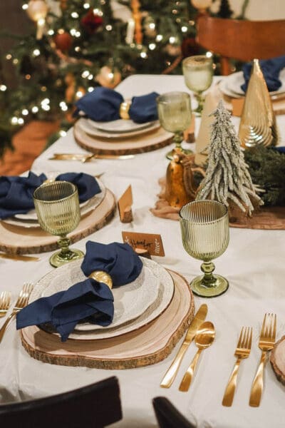 Christmas place setting with a navy napkin in a gold ring on cream beaded plates, a wood slice charger, gold flatware, and a green ribbed goblet beside a flocked mini tree