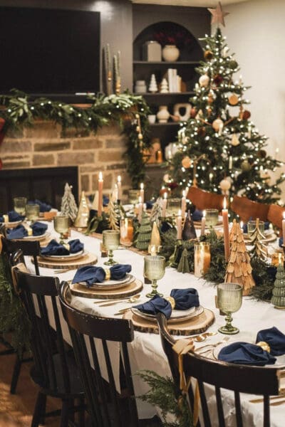 Long Christmas table with a white tablecloth, wood slice chargers, navy napkins in gold rings, green goblets, pine garland, and small trees in mixed shapes and materials, classic winter seasonal tablescape ideas