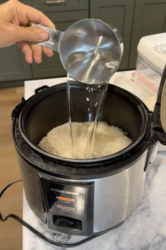 Crystel pouring measured water over rice in a rice cooker for Greek lemon chicken rice bowls