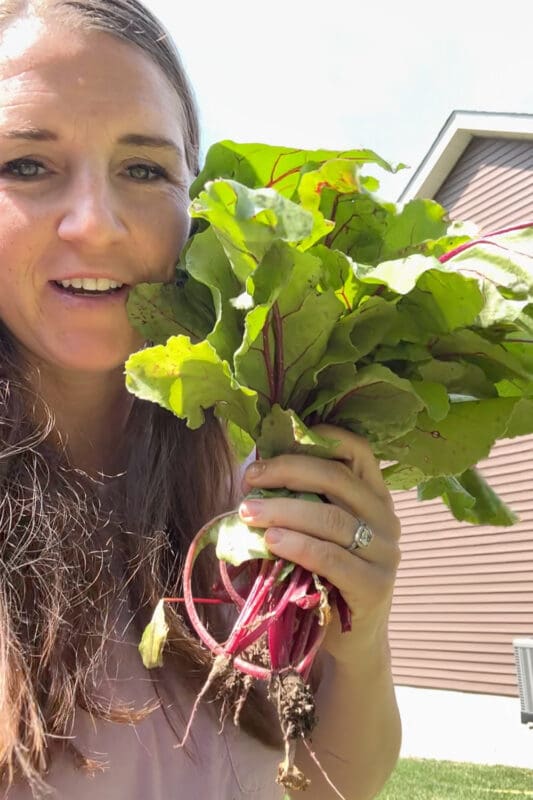 Crystel smiling and holding a freshly pulled bunch of beets with soil still on the roots and big leafy greens attached