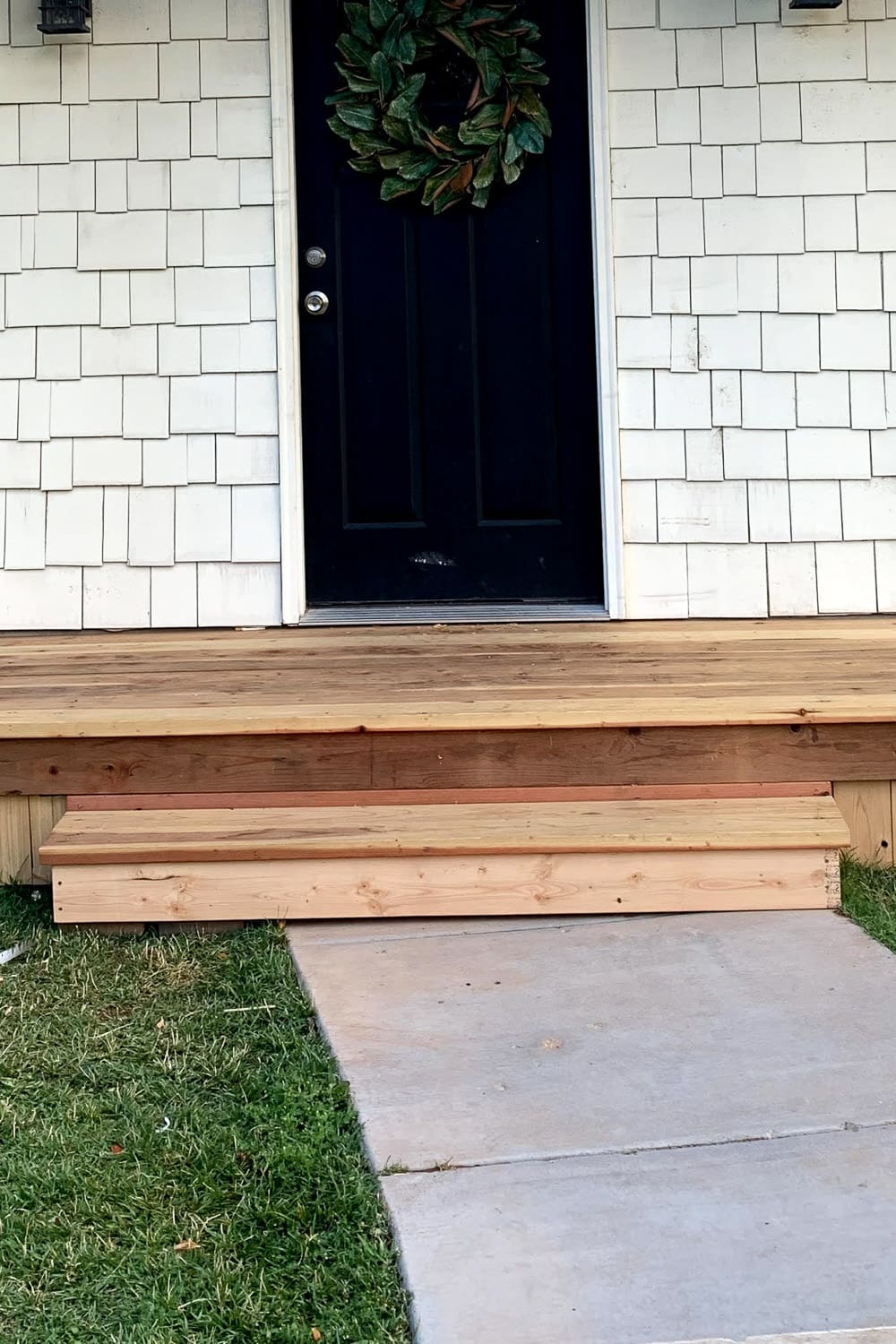 Close-up of the finished redwood porch step showing the tread overhang and skirted riser below a black front door with a magnolia wreath.
