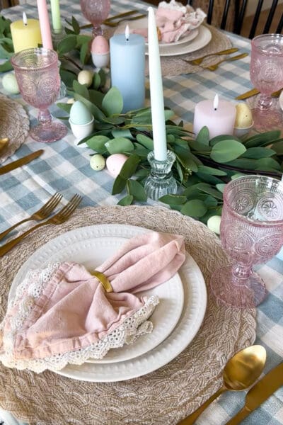 Easter place setting with a blush lace-trimmed napkin in a gold ring on white beaded plates, gold flatware, a pink pressed-glass goblet, and a round woven placemat