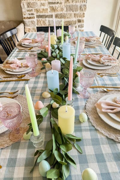 Easter table with a blue and cream gingham tablecloth, eucalyptus garland, pastel taper and pillar candles, and speckled eggs in white egg cups, fresh spring seasonal tablescape ideas