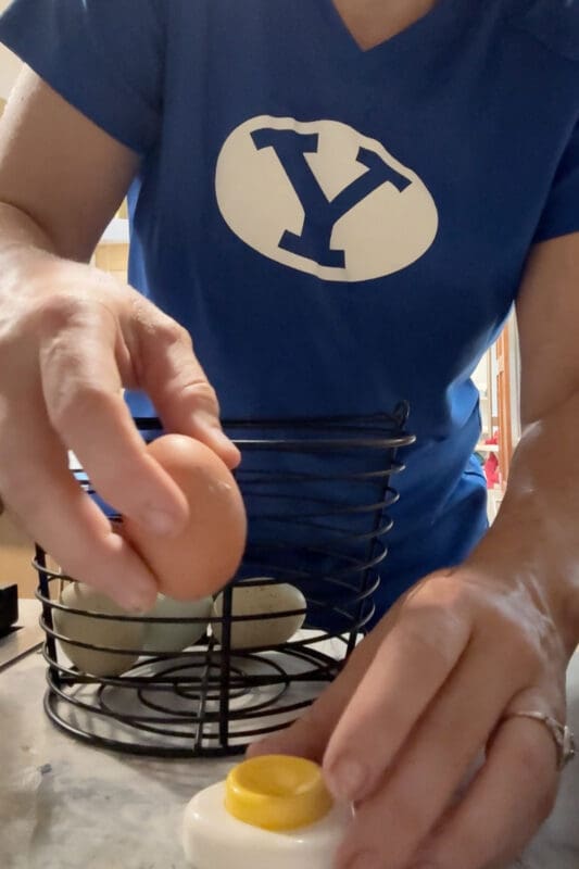 Crystel taking a fresh brown egg from a wire basket while pressing another egg onto an egg piercer, a small tool that punches a tiny hole in the shell before hard boiling fresh eggs so they peel easily.