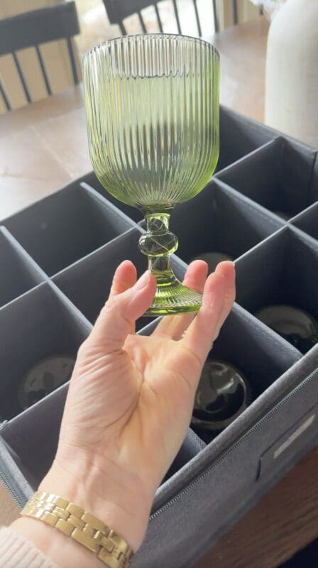 Hand lifting a green ribbed goblet from a gray padded storage box with divided compartments holding the rest of the set