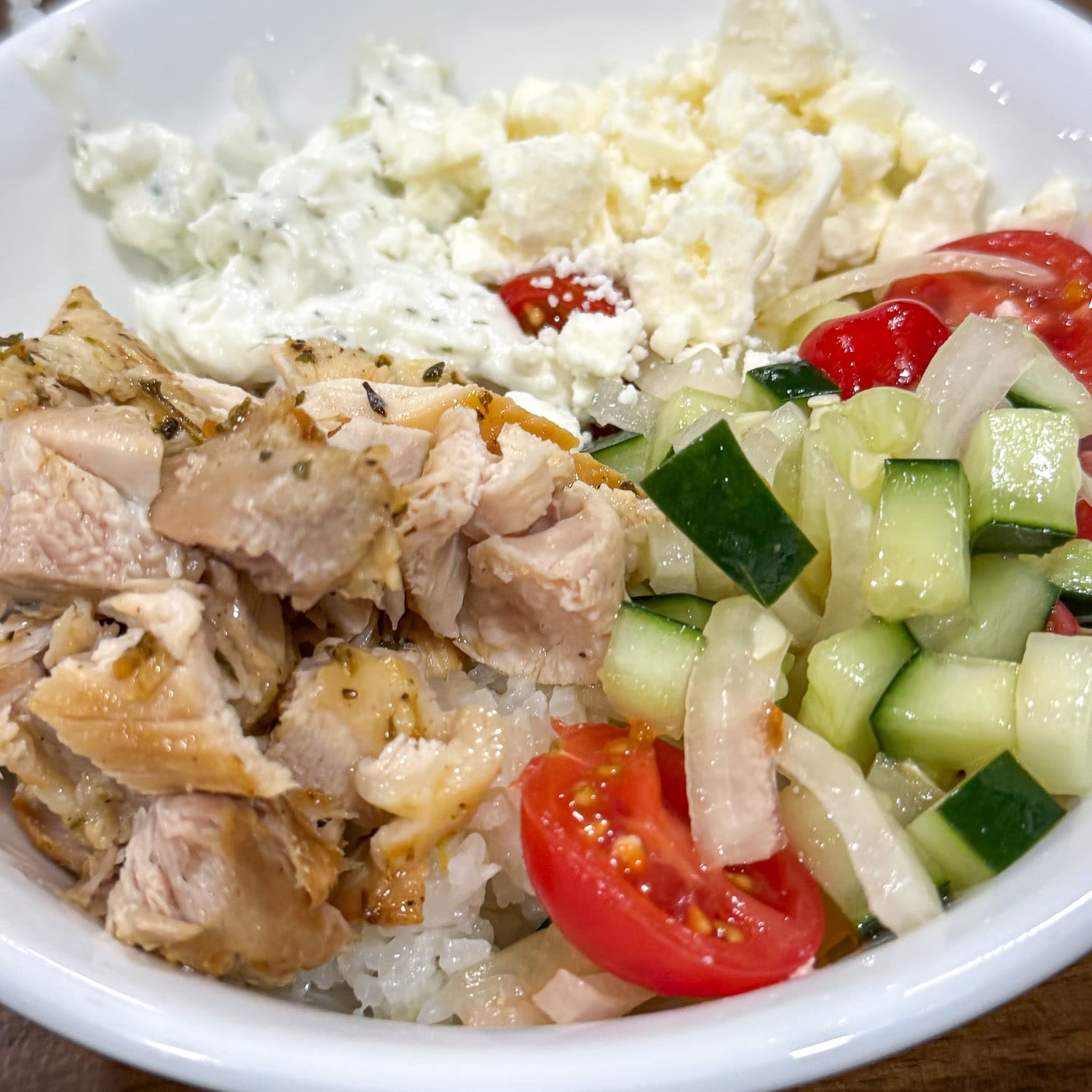 Close-up of Greek lemon chicken rice bowls showing sliced chicken over seasoned rice with crisp cucumber, cherry tomatoes, feta, and cool tzatziki
