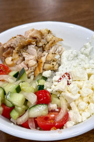 Greek lemon chicken rice bowls in a white bowl with chopped lemon-oregano chicken, cucumber-tomato salad, white onion, crumbled feta, and a scoop of tzatziki