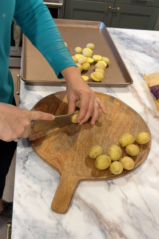 Crystel halving baby potatoes on a wooden cutting board with a sheet pan of cut potatoes nearby.