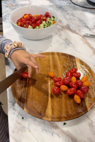 A young helper halving cherry tomatoes on a wooden board for the salad in the Greek lemon chicken rice bowls