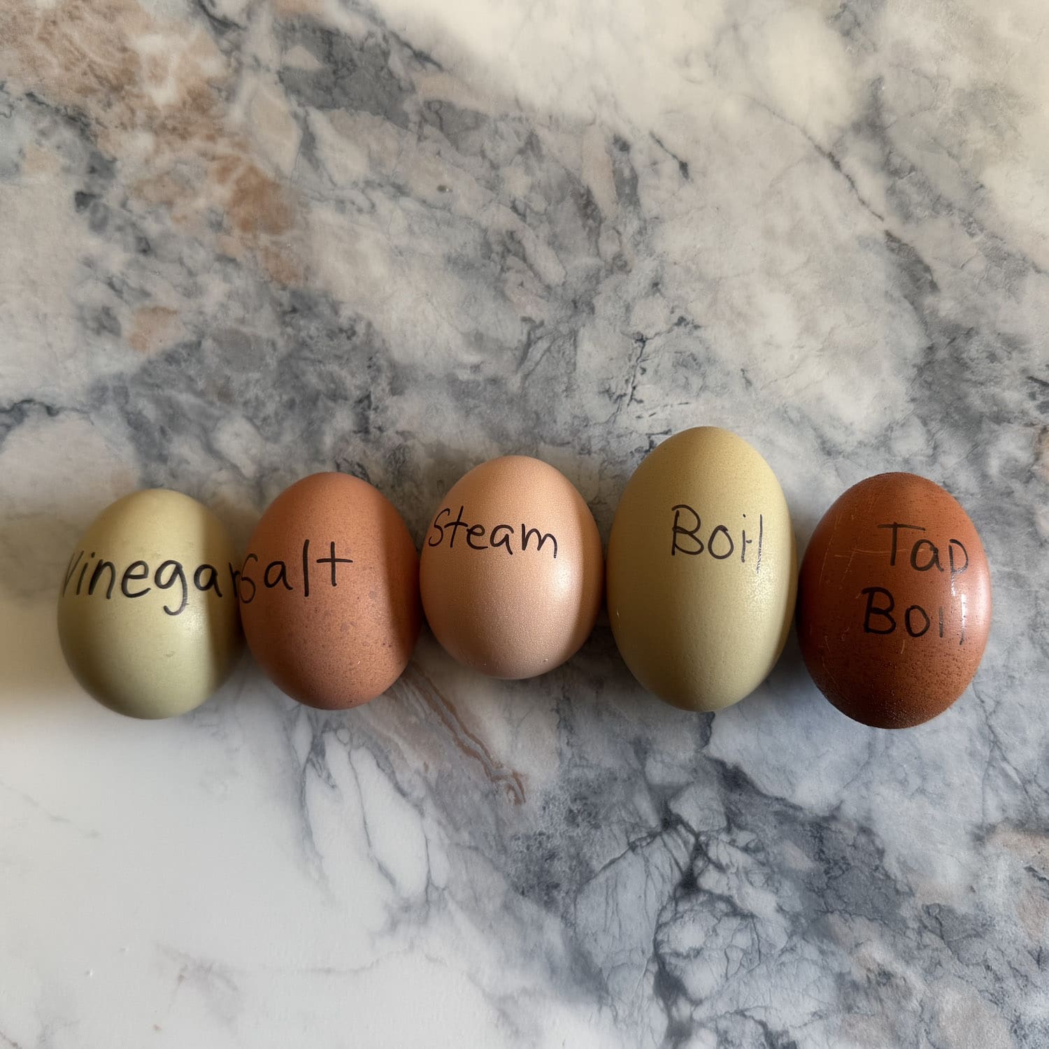 Close up of the same five labeled eggs on grey and white marble, comparing the vinegar, salt, steam, boil, and tap boil methods for hard boiling fresh eggs so they peel easily. Two small notes since these are near-duplicate framings: I gave them different file names and different alt phrasing so Google doesn't see them as redundant, and I kept the labeled-method list in both because that detail is genuinely what the image shows, which is good for accessibility and helps the image itself potentially rank for the method searches. Send the next batch whenever you're ready.