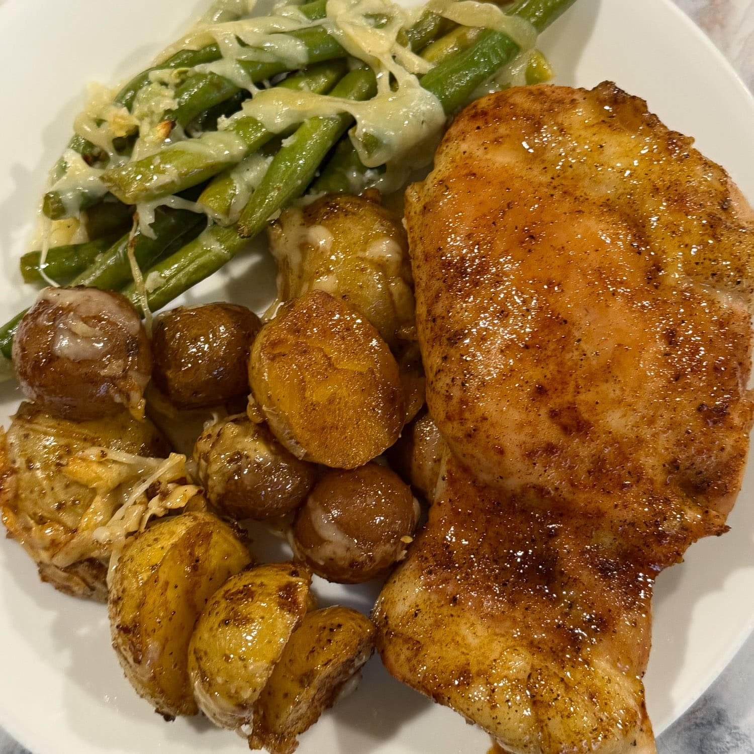 Close-up of a honey glazed sheet pan chicken thigh on a white plate with roasted baby potatoes and parmesan green beans.