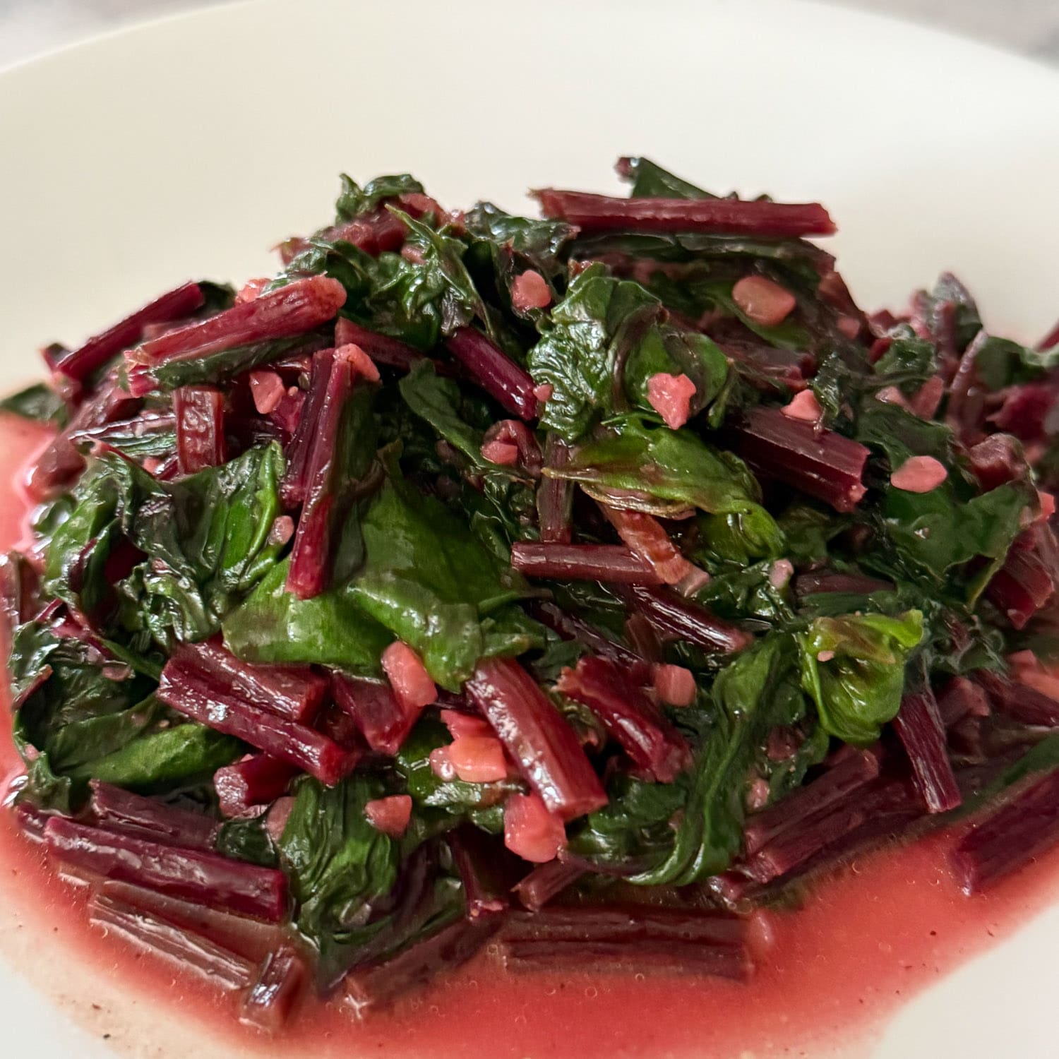 Crystel's cooked beet greens with tender leaves and ruby stems on a white plate with pink pot liquor pooling around them