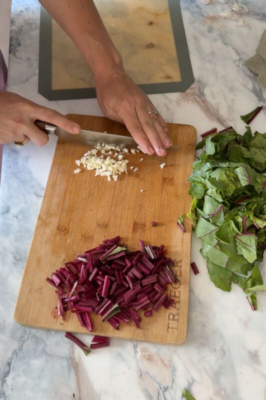 Crystel finely mincing garlic on a Traeger board with chopped beet green stems and leaves waiting alongside