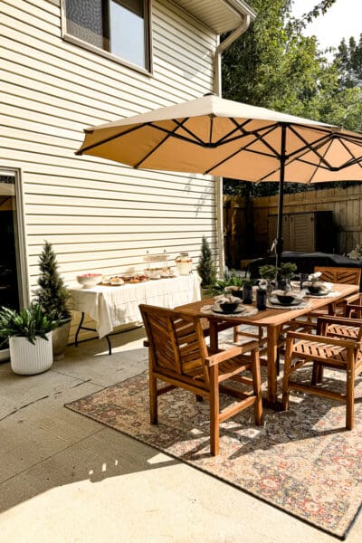 Backyard patio set for outdoor entertaining with a wood dining table under a tan umbrella, woven placemats, and a dessert table set up along the house